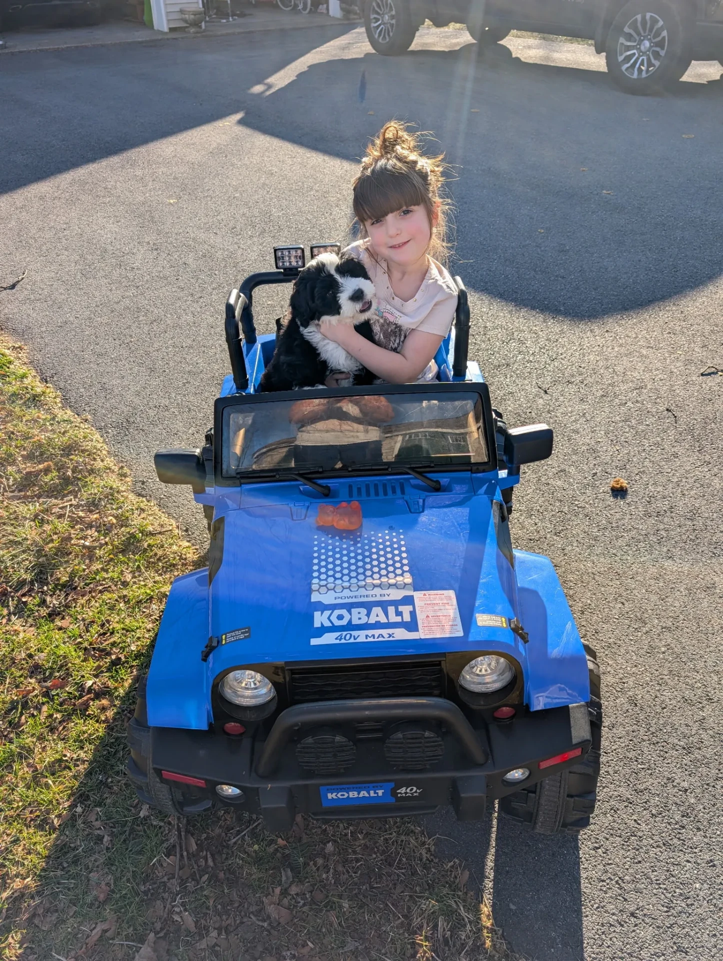 A girl holding a black and white puppy in a blue toy Jeep in the driveway