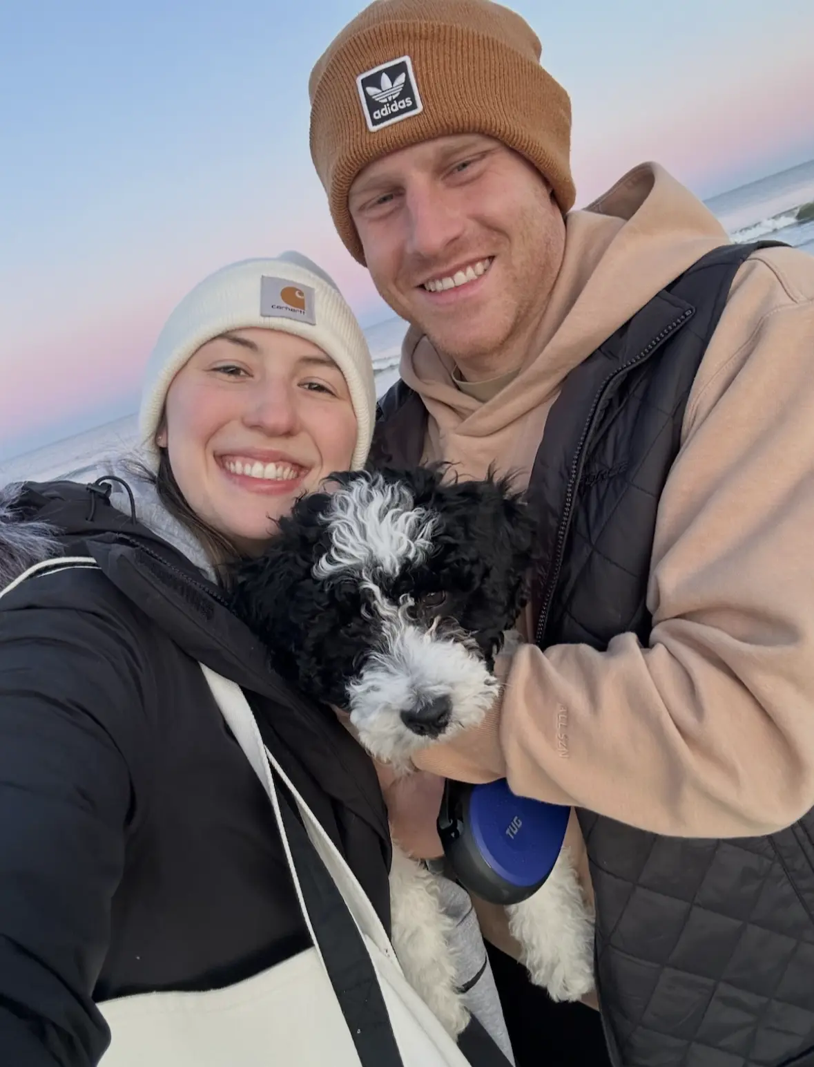 A couple in winter hats holding their black and white puppy at the beach